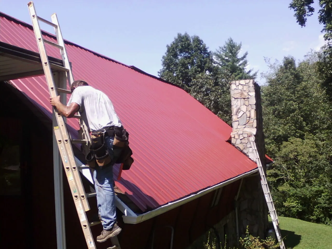 Roofer on a ladder beside a red metal roof and stone chimney — Soffit & Fascia Repair by Roofing Brothers in Shasta